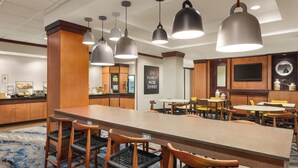 A hotel lobby with a long communal table, several chairs, hanging pendant lights, and a buffet area in the background.