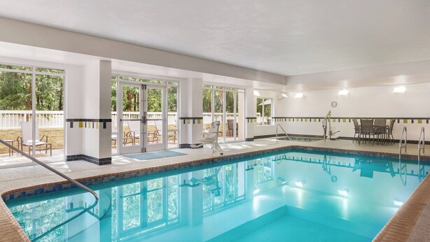 A large indoor swimming pool with clear glass walls, a tiled deck area, and a poolside seating arrangement.
