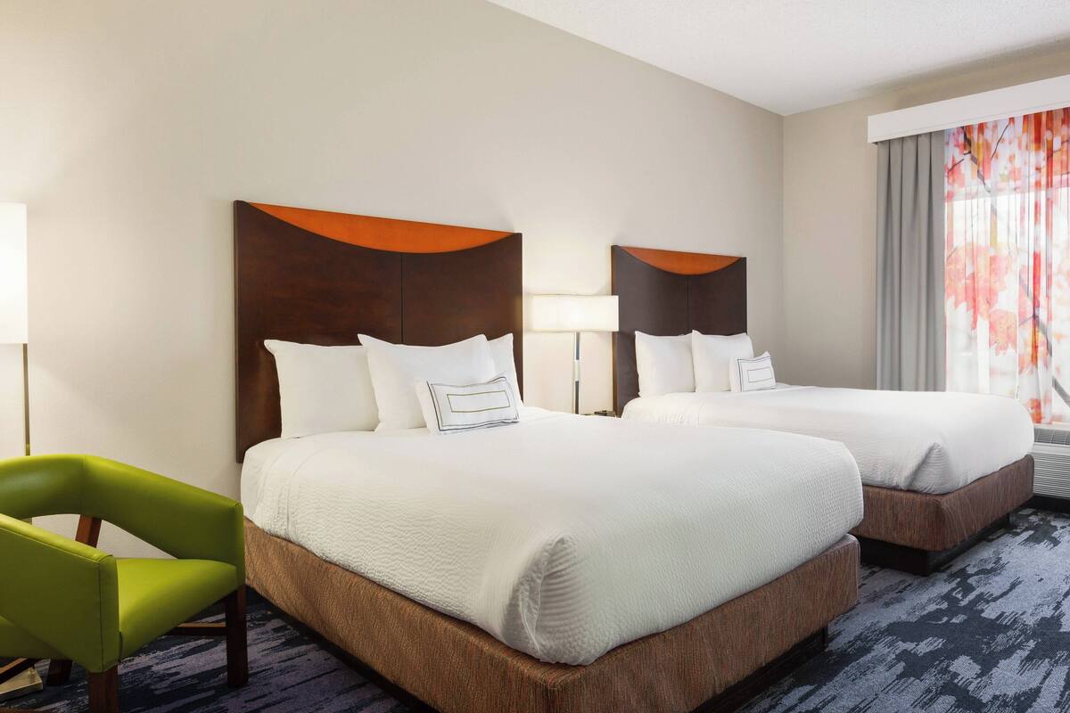 Habitación de hotel con dos camas, una silla verde y un ventanal con cortinas.