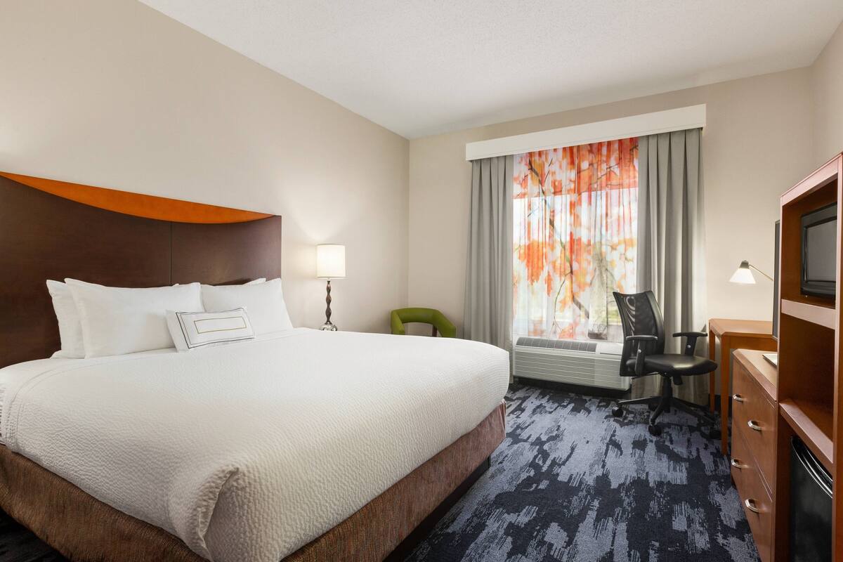 Una habitación de hotel con una cama grande, un escritorio, una silla, un televisor y un ventanal con cortinas.