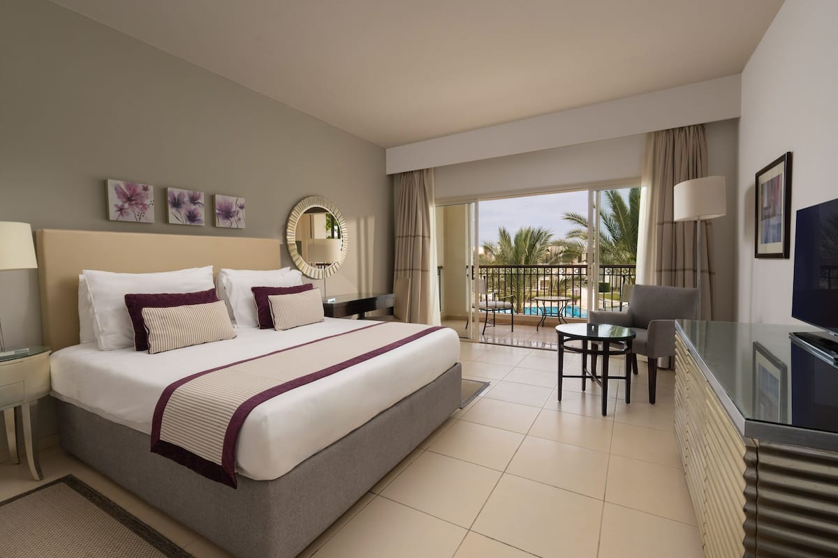 A hotel room with a large bed, a desk, a chair, a TV, and a view of a pool and palm trees.