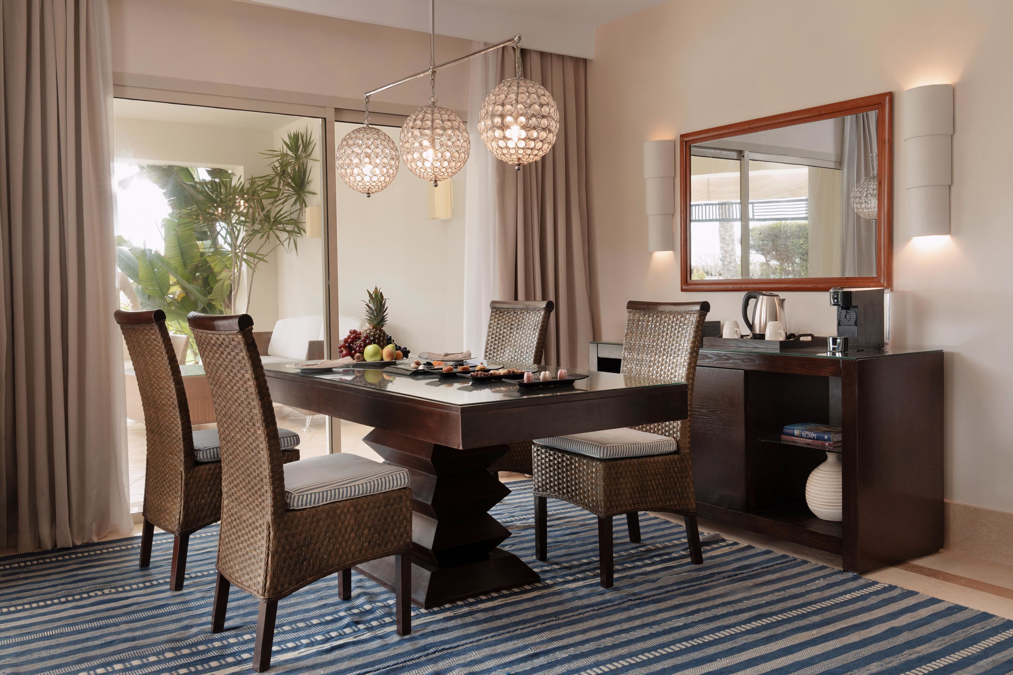 A dining area with a large table, wicker chairs, a chandelier, a mirror, and a console table with a vase.