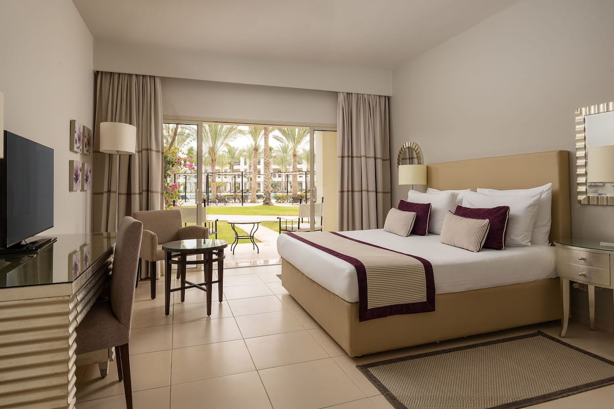 A hotel room with a bed, desk, TV, and a view of a courtyard with palm trees.
