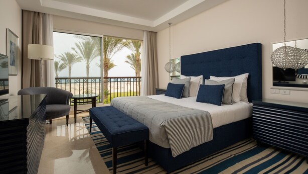 A modern hotel room with a large bed, a seating area, and a balcony with a view of palm trees.