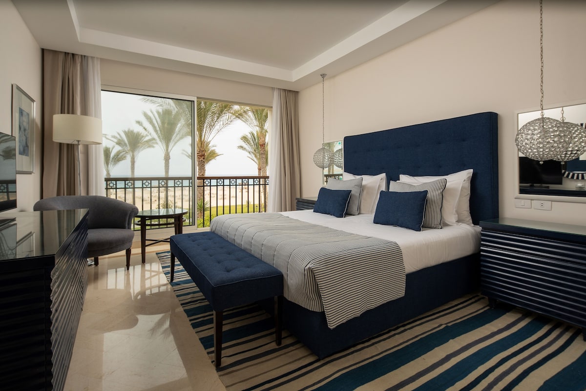 A modern hotel room with a large bed, a seating area, and a balcony with a view of palm trees.