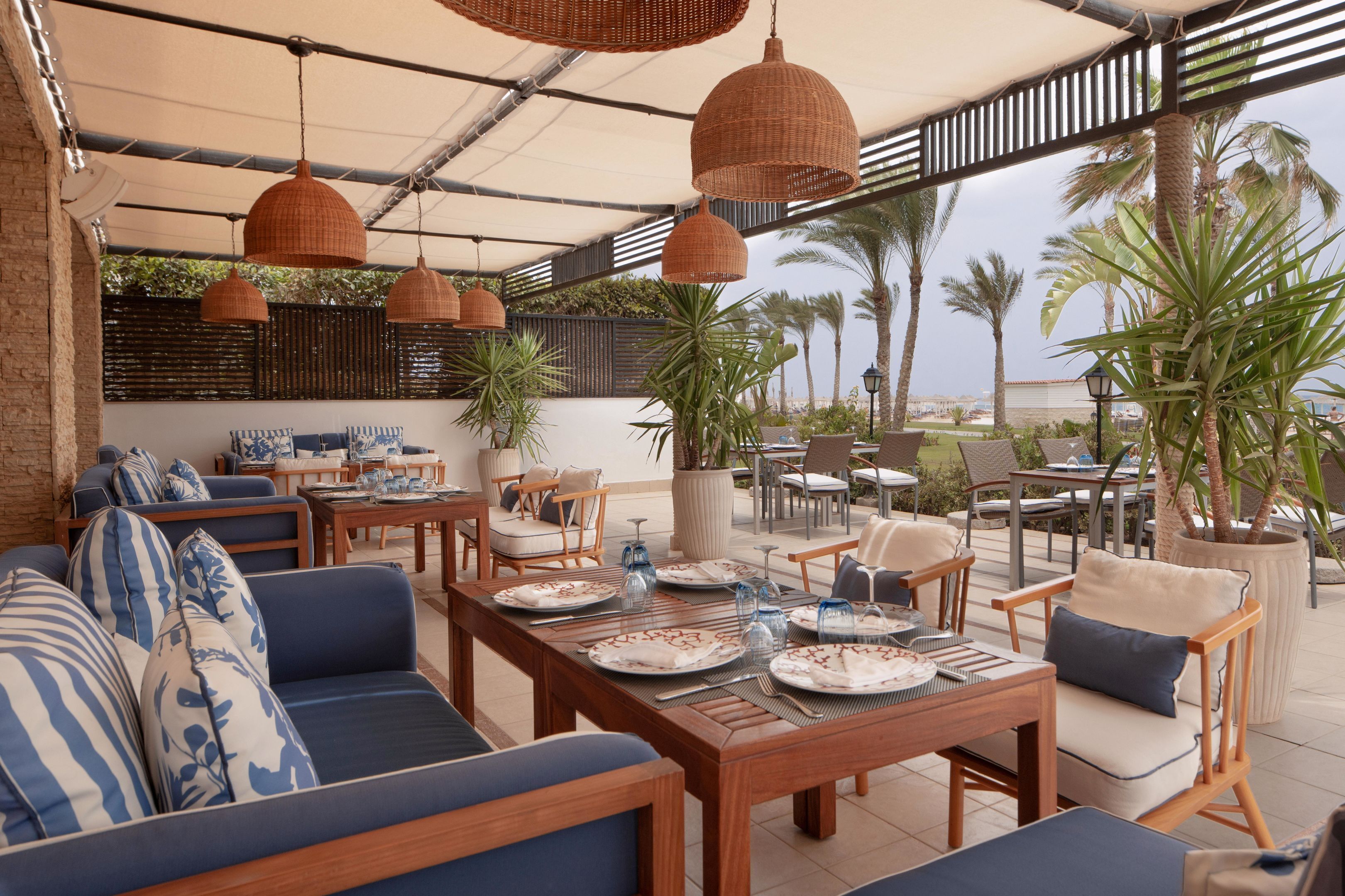 An outdoor dining area with wooden tables and chairs, blue and white patterned cushions, hanging wicker lamps, and a view of palm trees and the sea.