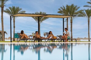 A poolside area with lounge chairs and a canopy, palm trees, and a clear sky.