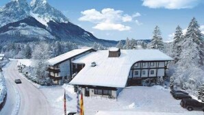 A snow-covered building with a sloped roof, surrounded by snow-laden trees and mountains in the background.