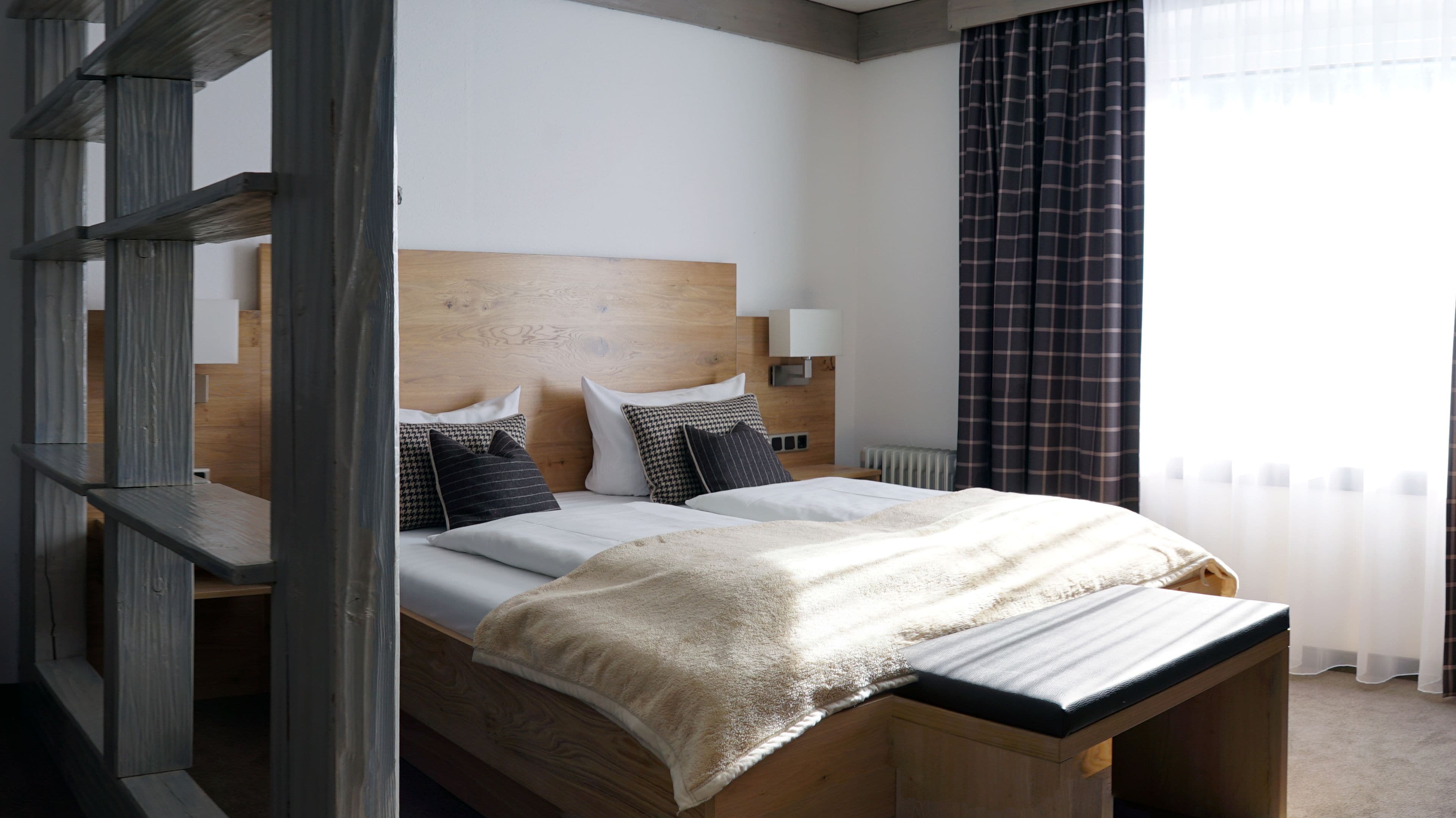 A bedroom with a wooden headboard, a bed with white bedding and multiple pillows, a bedside table, and a window with curtains.