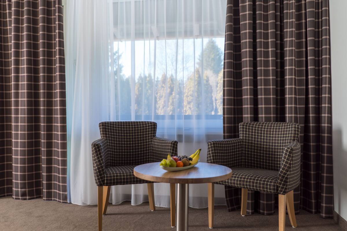 Ein Zimmer mit zwei karierten Sesseln, einem runden Tisch mit Obstschale und transparenten Vorhängen mit Blick auf Bäume.