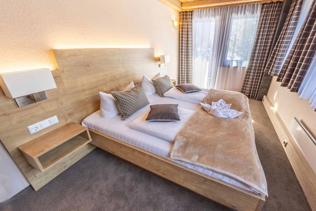 Ein Zimmer mit einem Einzelbett, einem Holzkopfende, Nachttischen, einer eingebauten Ablage und einem Fenster mit Vorhängen.