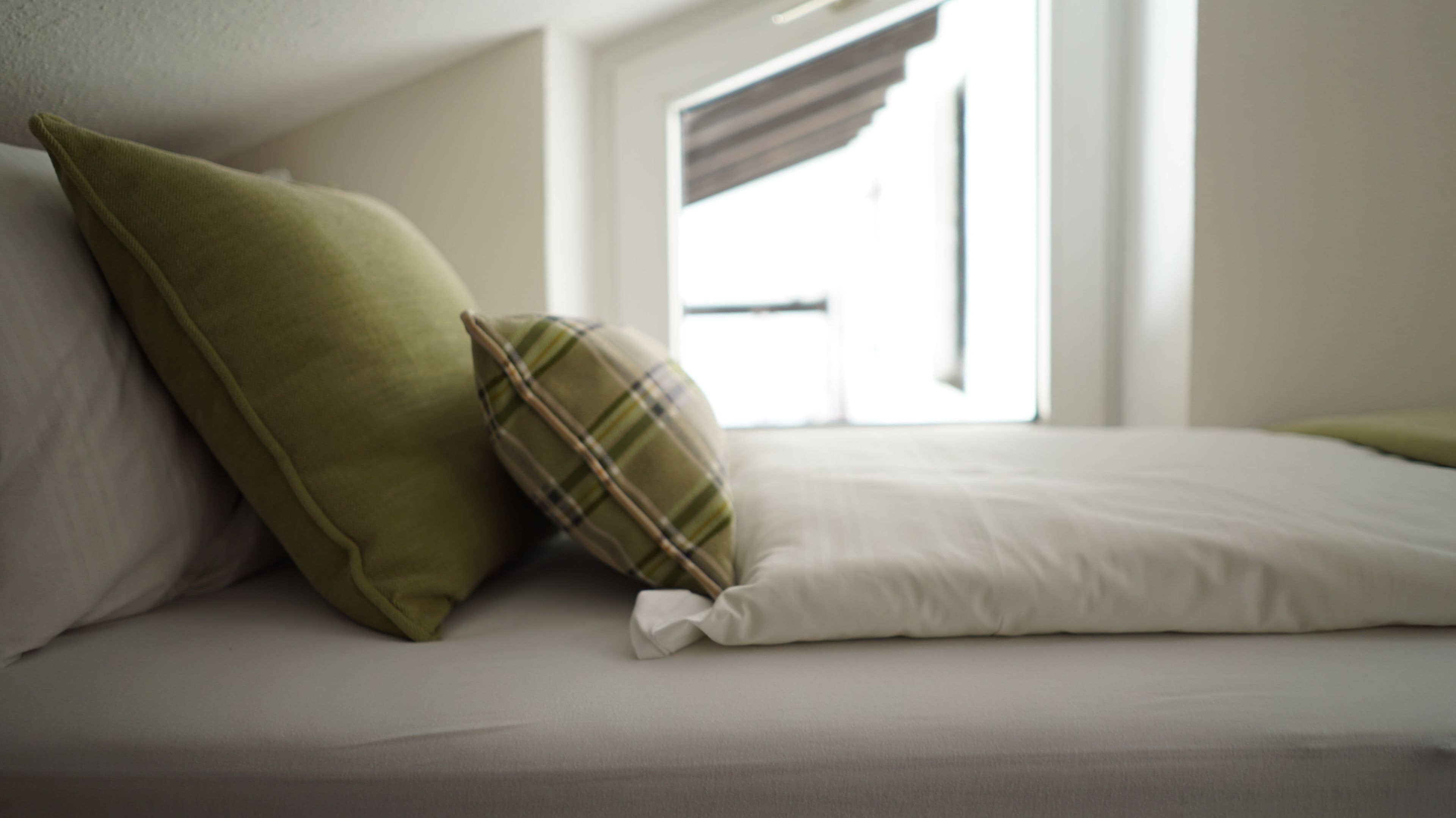Ein Bett mit zwei Kissen, eines grün und eines kariert, in einem Zimmer mit Fenster und weißer Wand.