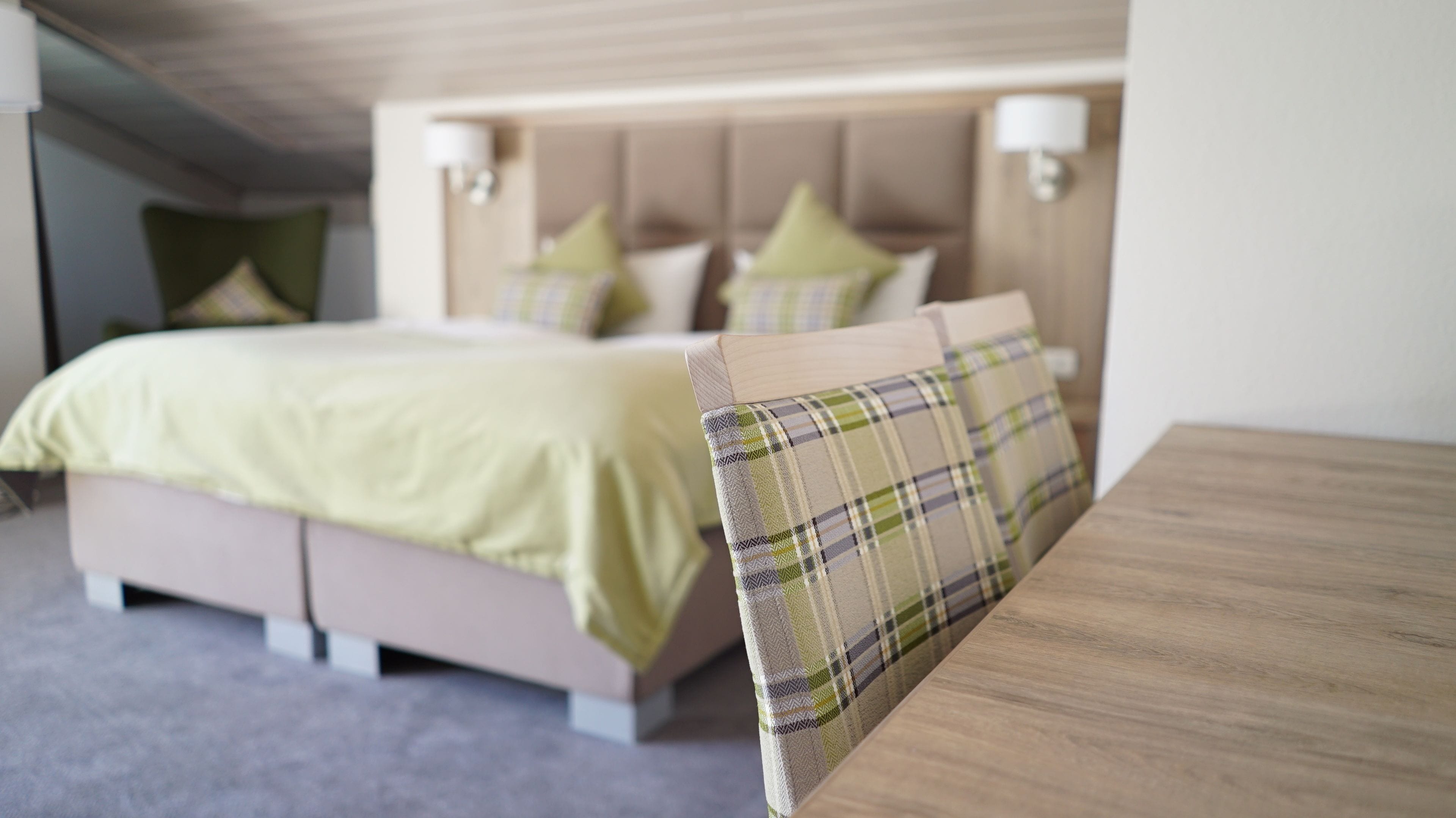 A neatly made bed with a green bedspread and matching pillows, a wooden headboard, and a bedside table with a plaid pillow.