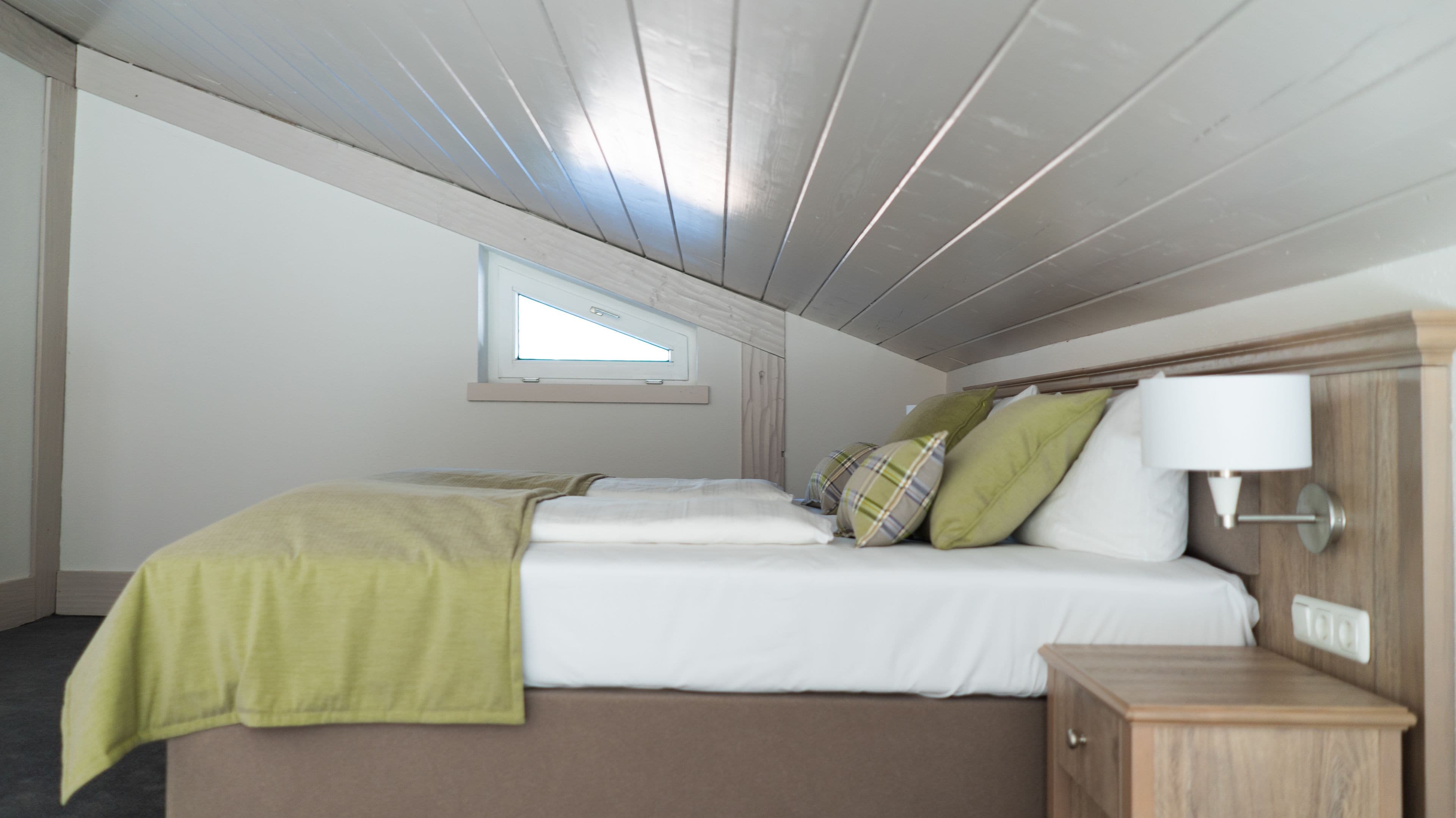 Un dormitorio con cama, almohadas, mesita de noche, lámpara y ventana.