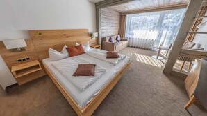 A modern hotel room with a large bed, a wooden headboard, a bedside table, a small table, and a view of the outdoors through large windows.