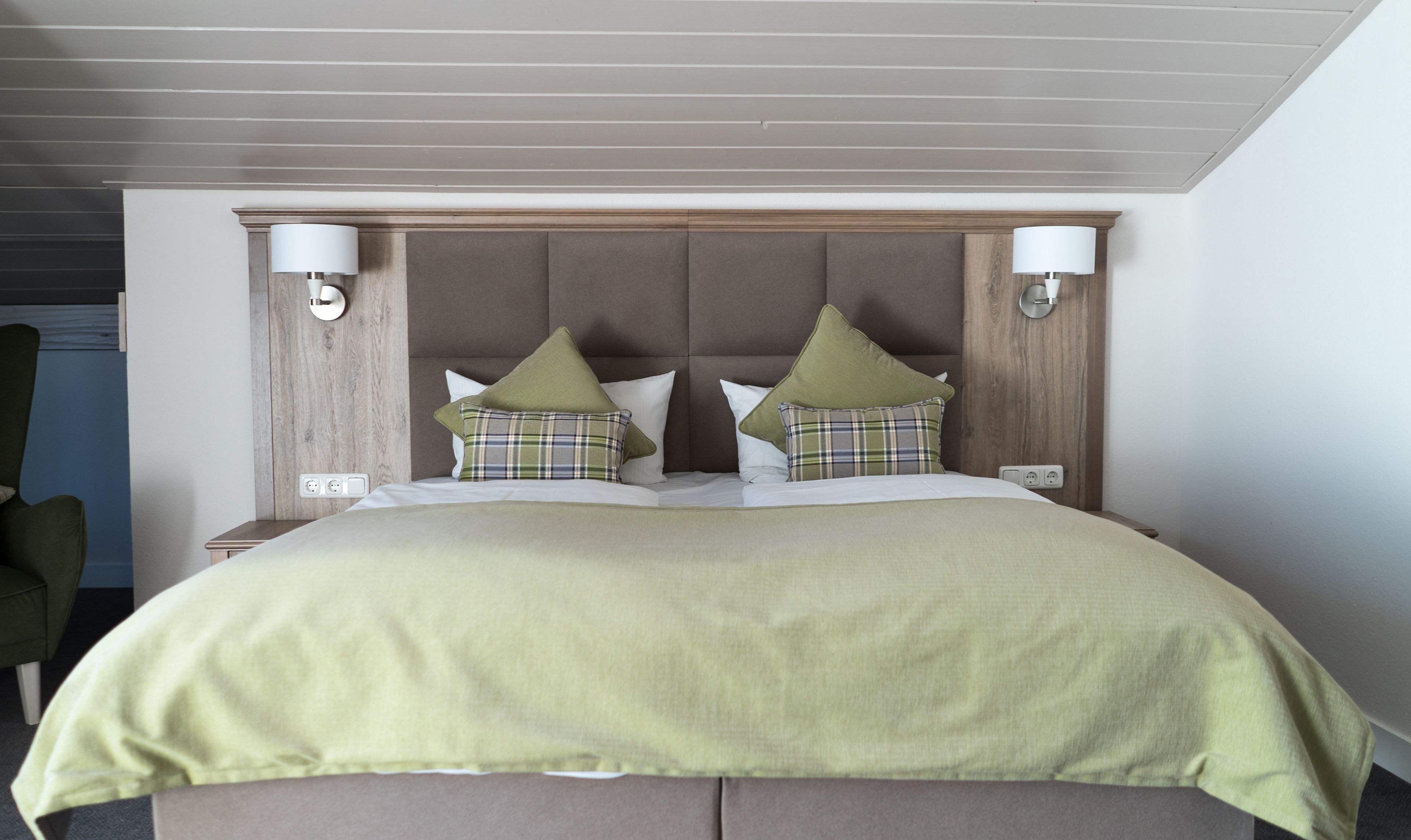 A hotel room with a bed, headboard, two wall-mounted lamps, and a green chair.