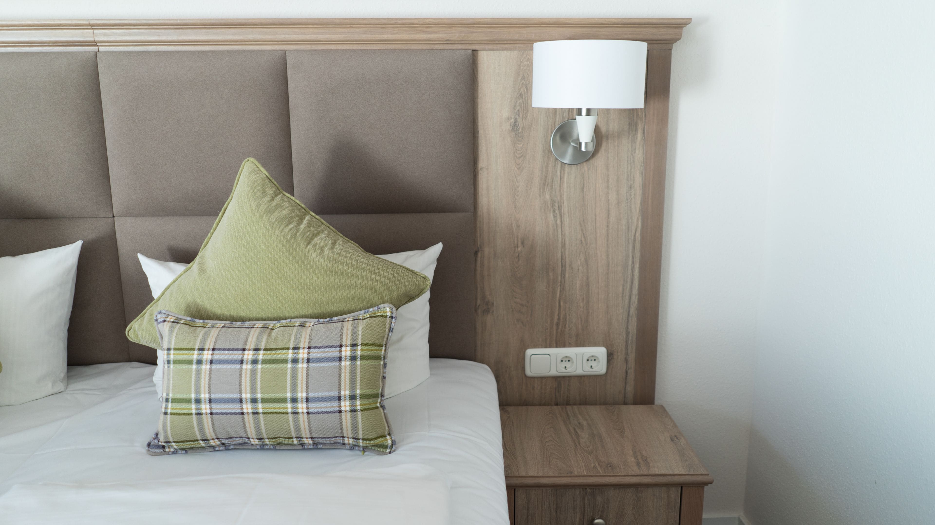 Una cama bien hecha con un cojín escocés y uno verde, una mesita de noche de madera con una lámpara, y un enchufe de pared.