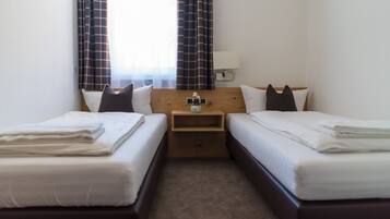 A hotel room with two single beds, a wooden headboard, a small bedside table with a vase, and a window with curtains.