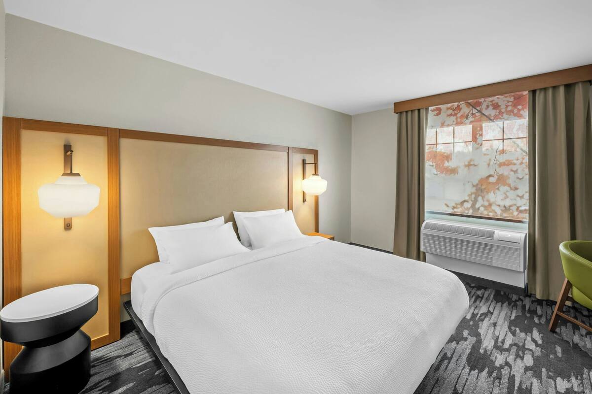 Une chambre d’hôtel avec un grand lit, deux lampes de chevet, une fenêtre avec des rideaux, un radiateur et un fauteuil vert.