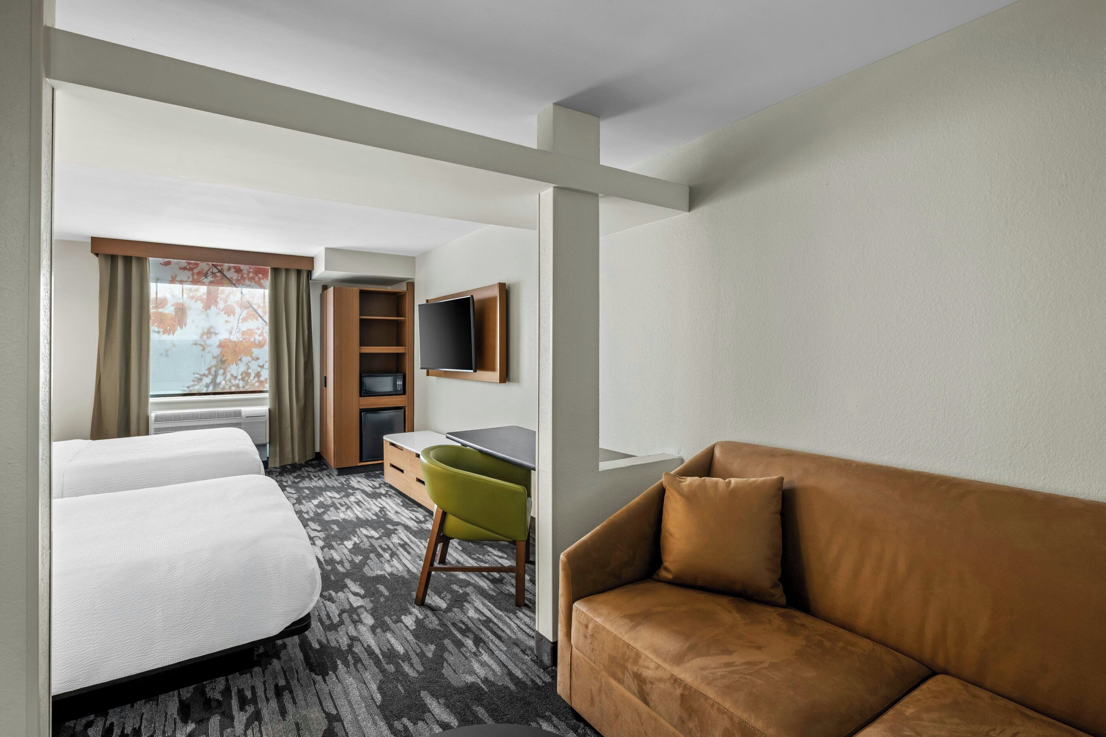 A hotel room with two beds, a desk, a green chair, a brown sofa, and a TV mounted on the wall.