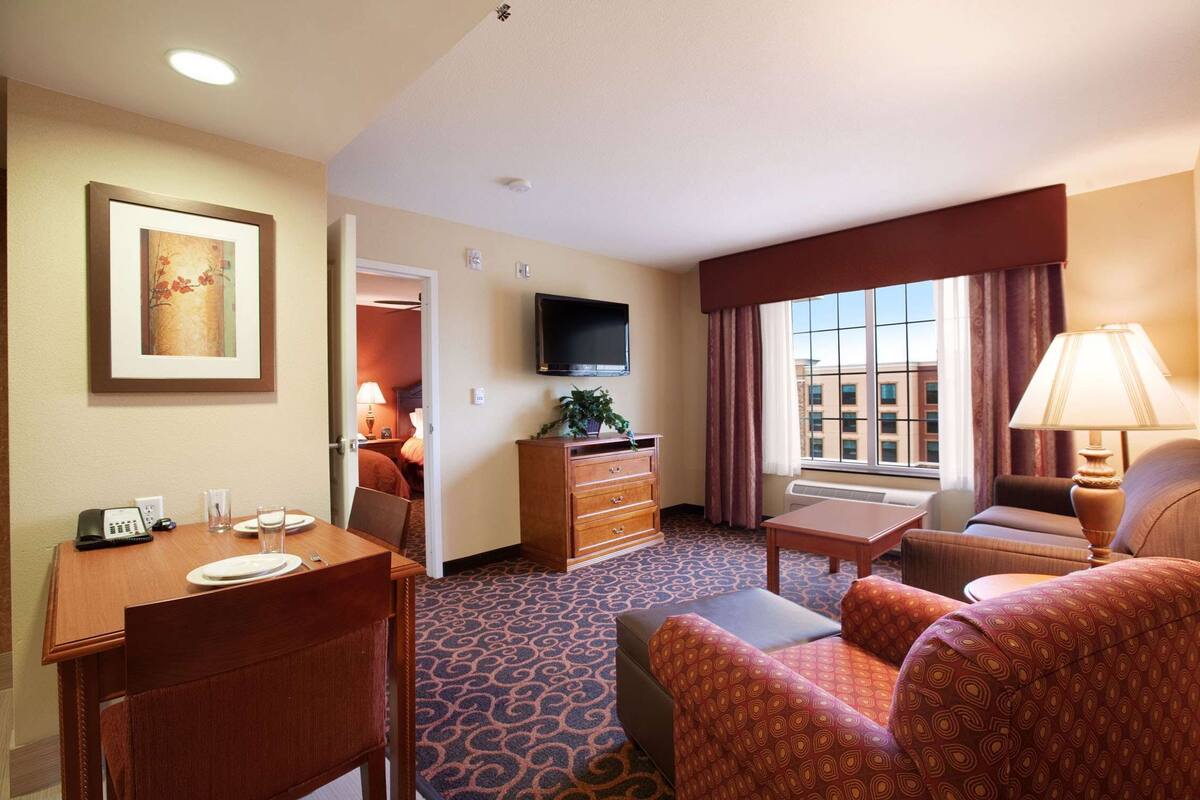 A hotel room with a dining table, a sofa, a TV, a dresser, and a window with curtains.