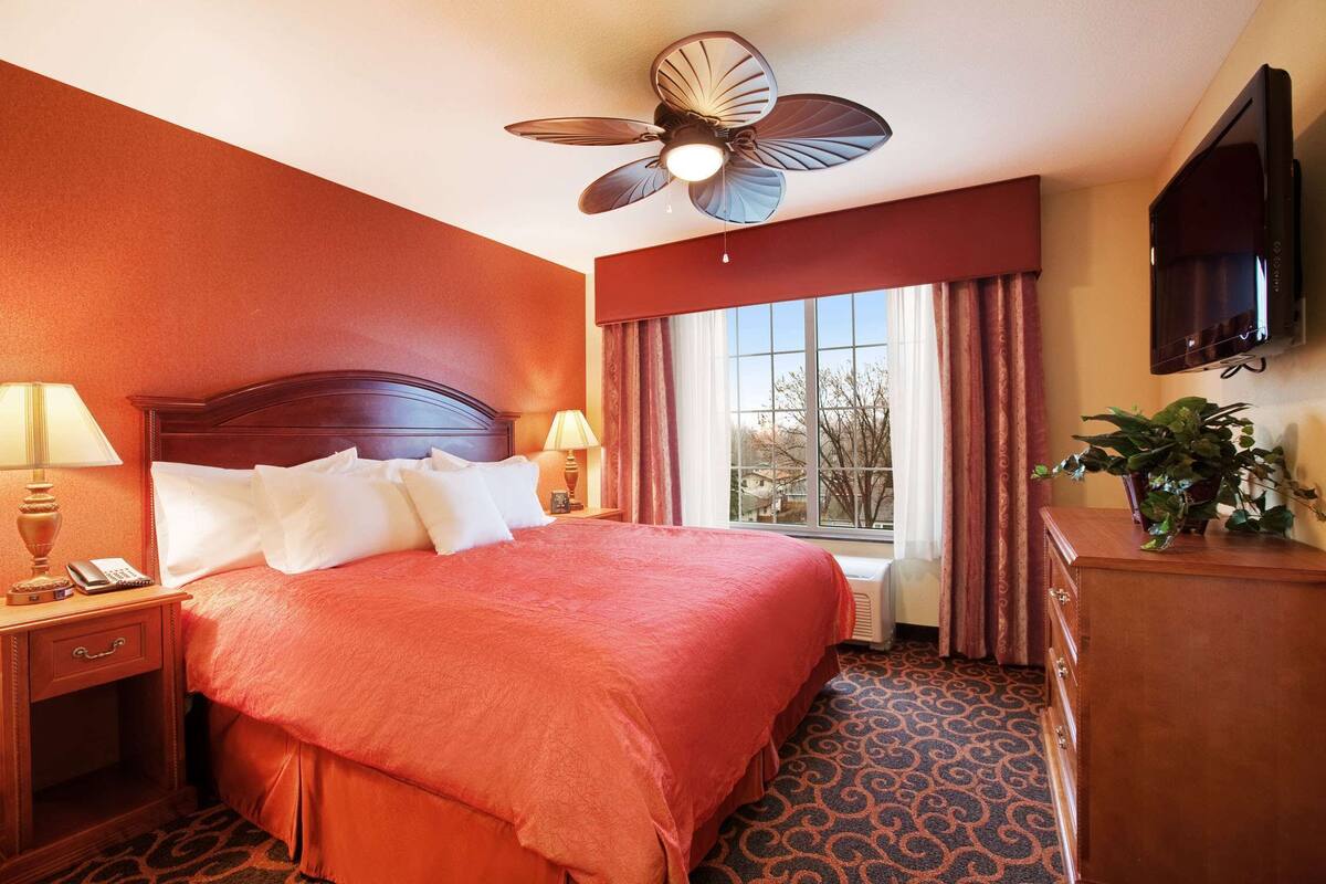 A hotel room with a large bed, a ceiling fan, a TV, and a window with curtains.