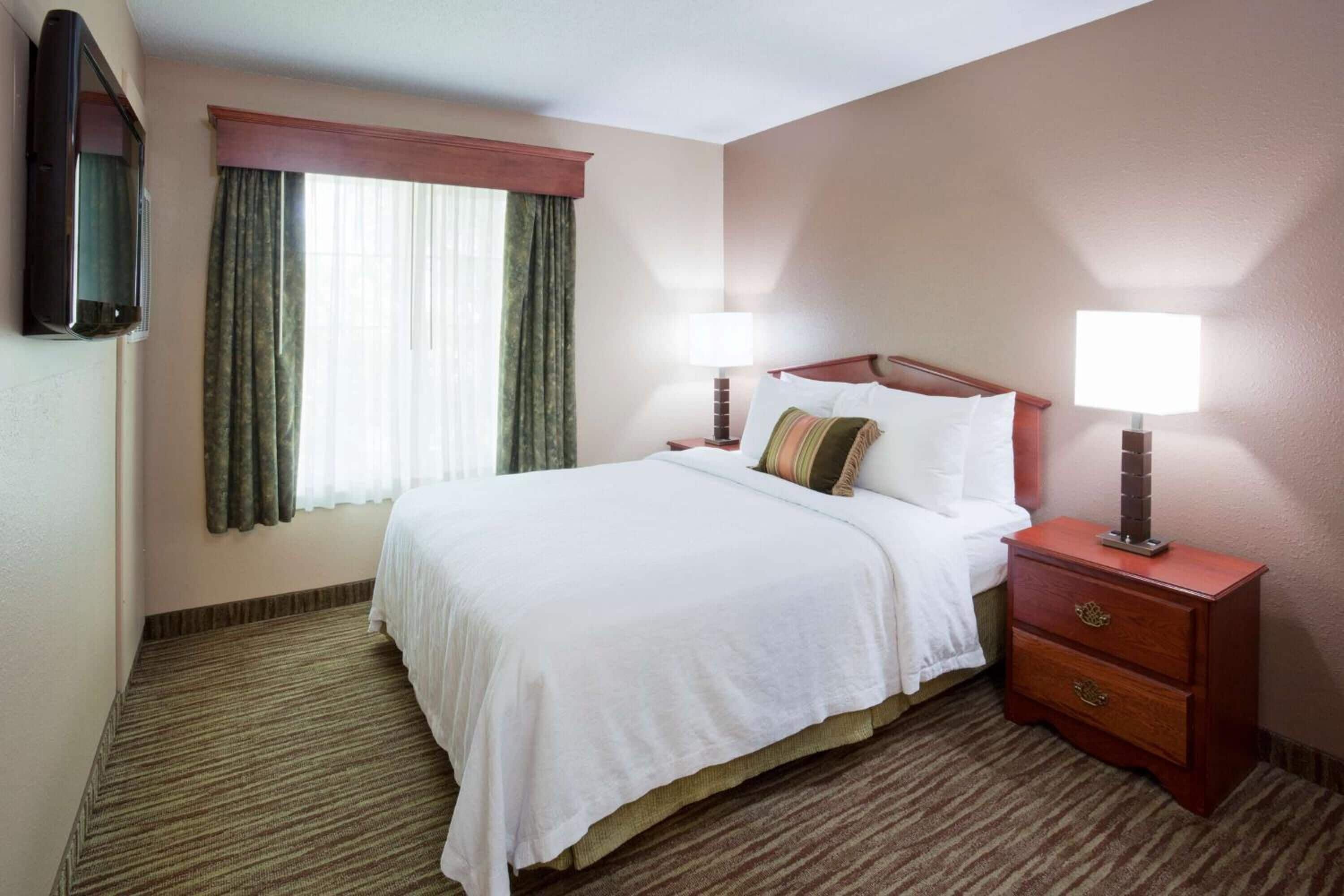 A hotel room with a single bed, two bedside tables with lamps, a flat-screen TV mounted on the wall, and a window with curtains.
