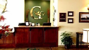 A reception desk with a green wall and a large 'G' logo. There are framed pictures on the wall and a potted plant to the right. - GrandStay Residential Suites Hotel (Faribault)