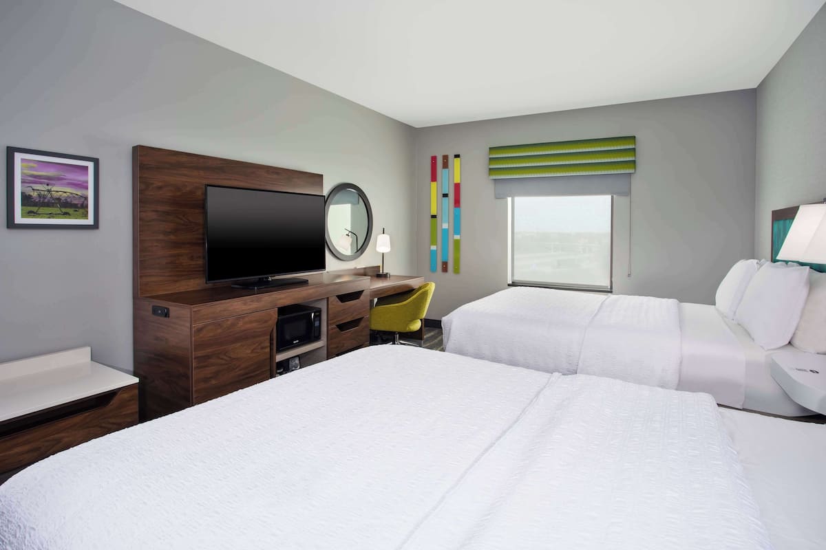 A hotel room with two beds, a TV, a desk with a chair, a window with a view, and a wall with a colorful artwork.