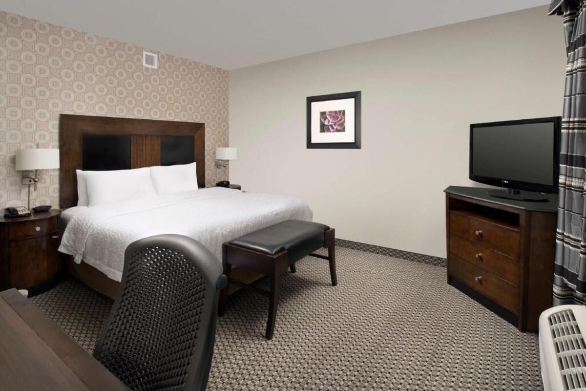 Quarto de hotel com cama, mesas de cabeceira, televisão, escrivaninha e uma cadeira.