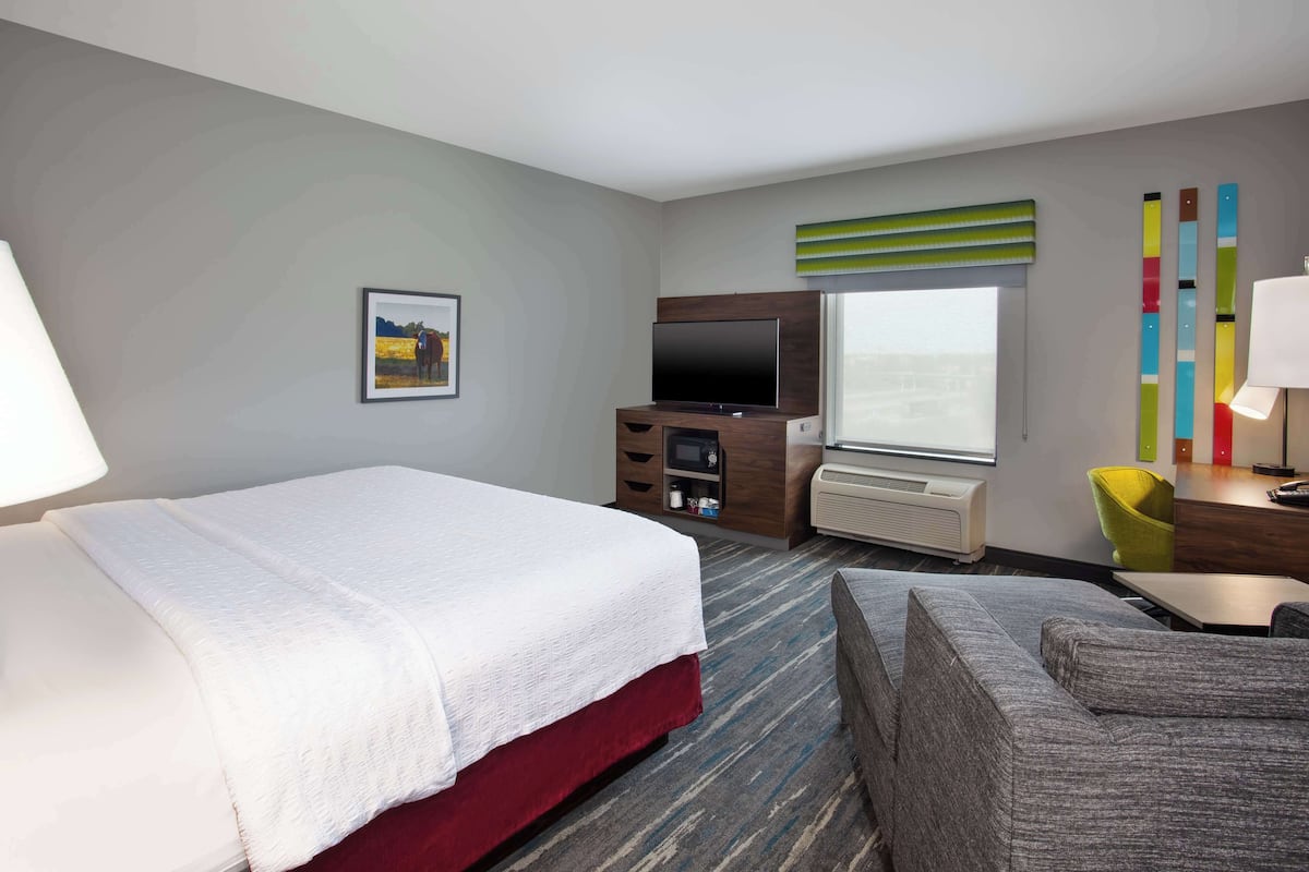 A hotel room with a bed, a TV, a desk with a lamp, a chair, and a window with blinds.