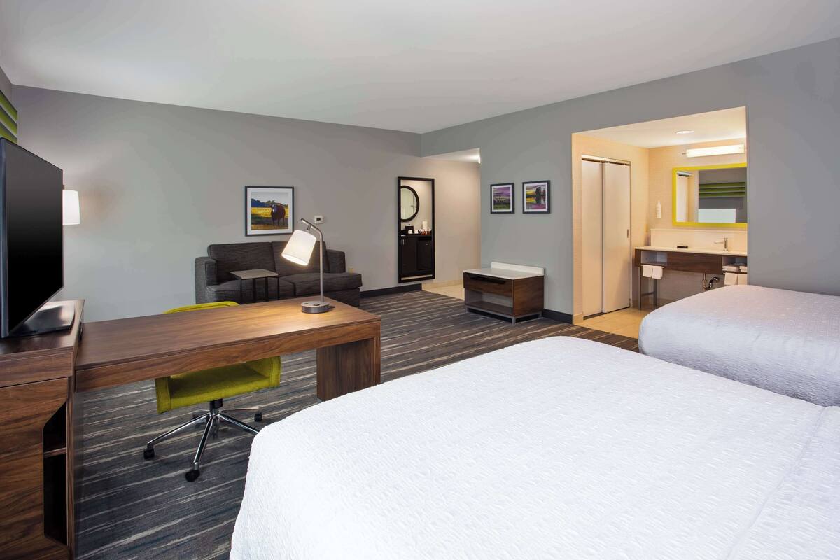 A hotel room with a bed, a desk, a chair, a TV, a lamp, and a bathroom with a mirror and sink.