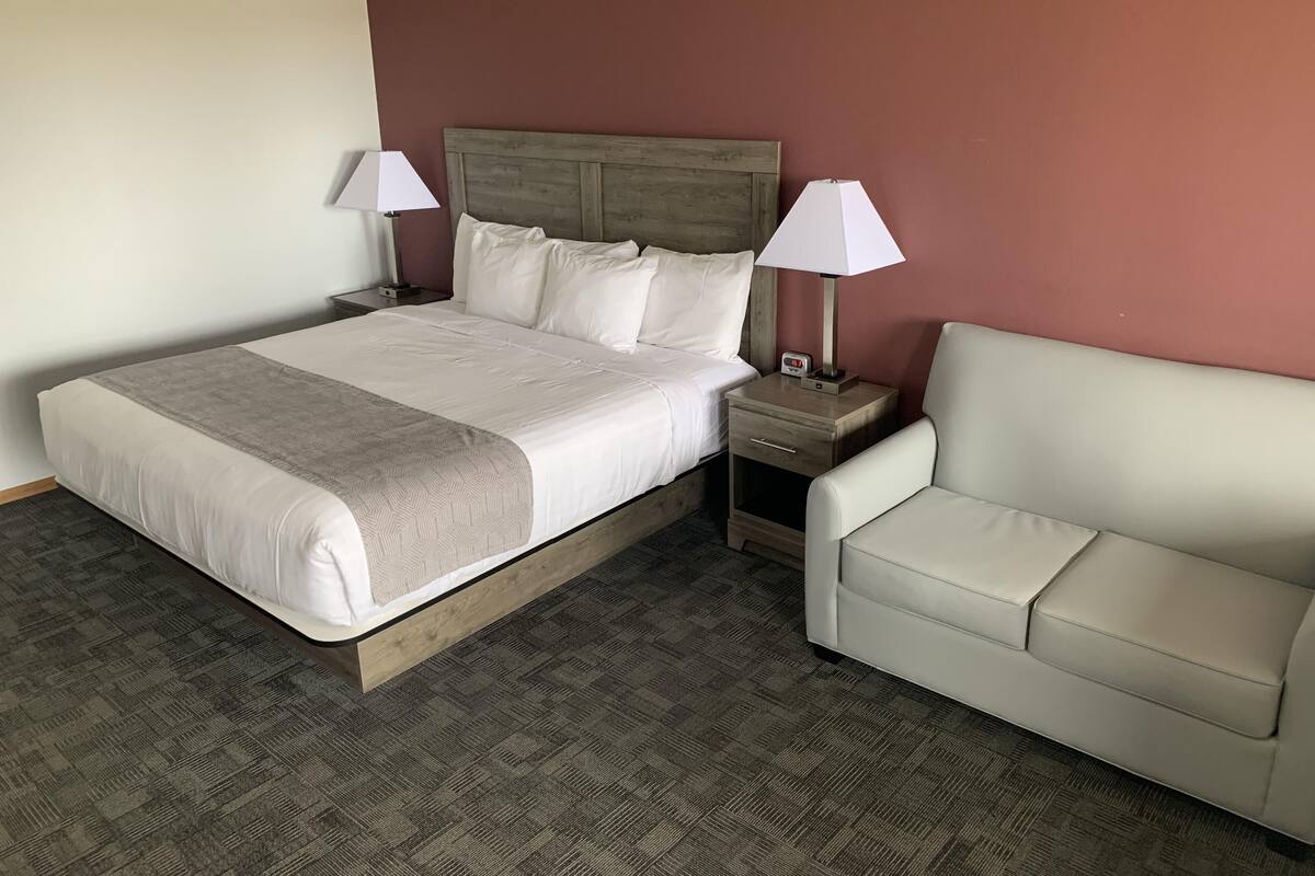 A hotel room with a bed, two bedside lamps, a nightstand, a white sofa, and a wooden headboard.