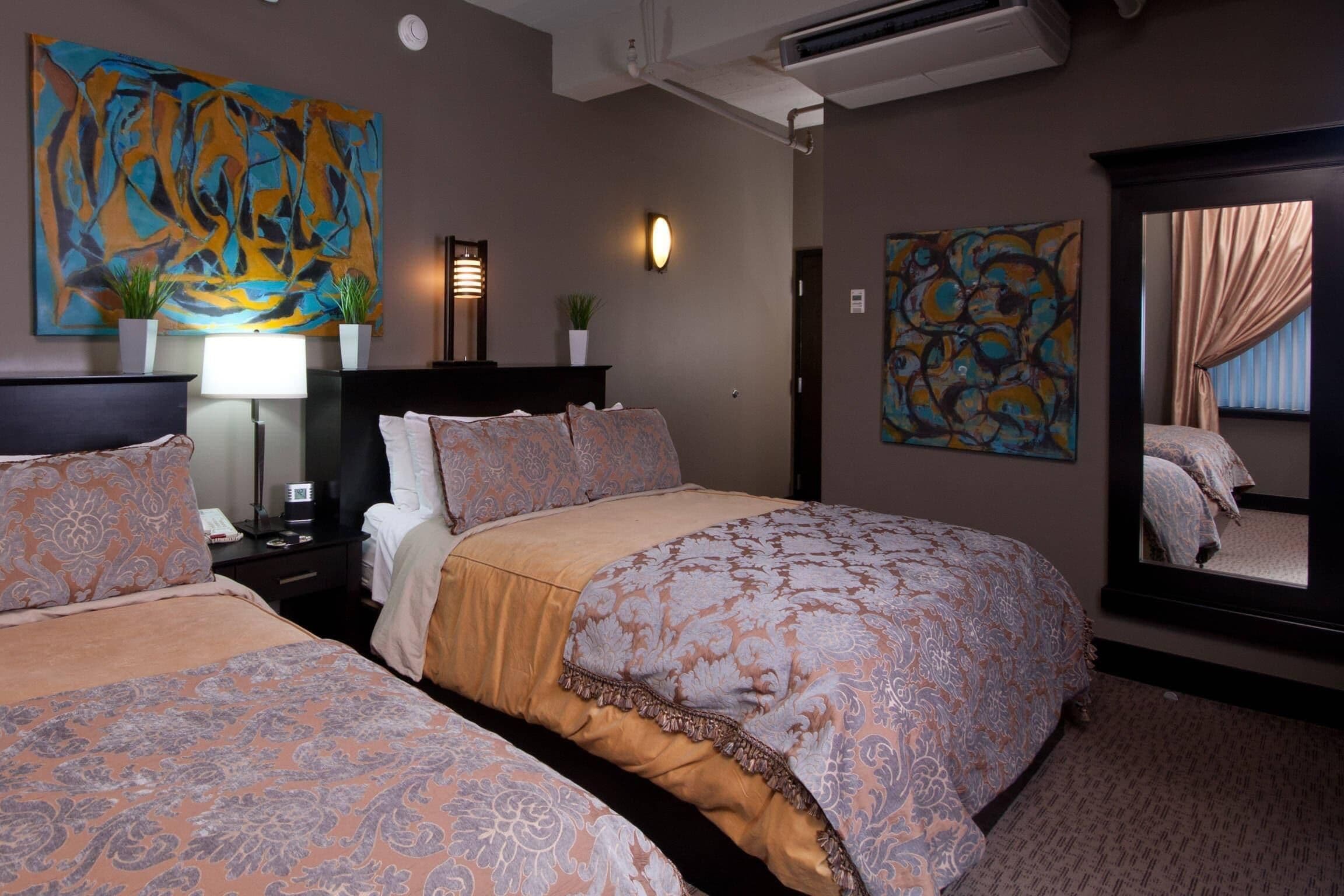 Habitación de hotel con dos camas, un cuadro abstracto grande, una lámpara de pared y un espejo que refleja la habitación.