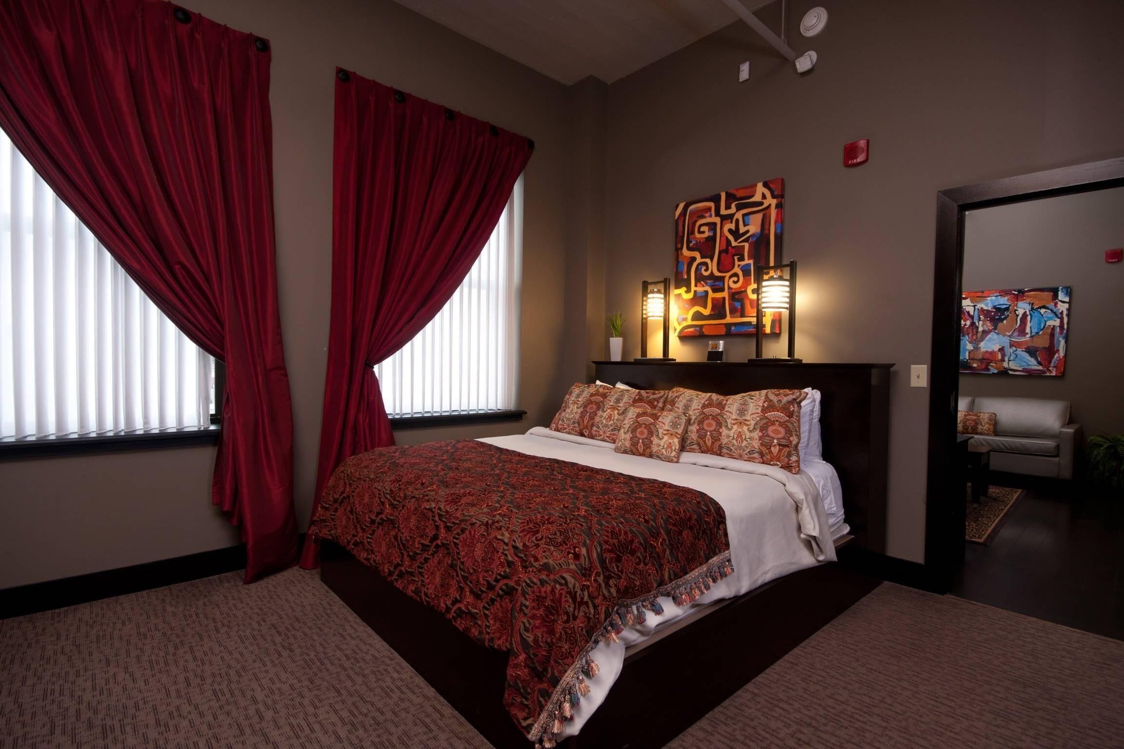 A bedroom with a large bed, red curtains, a wall-mounted artwork, and a side table with a lamp.