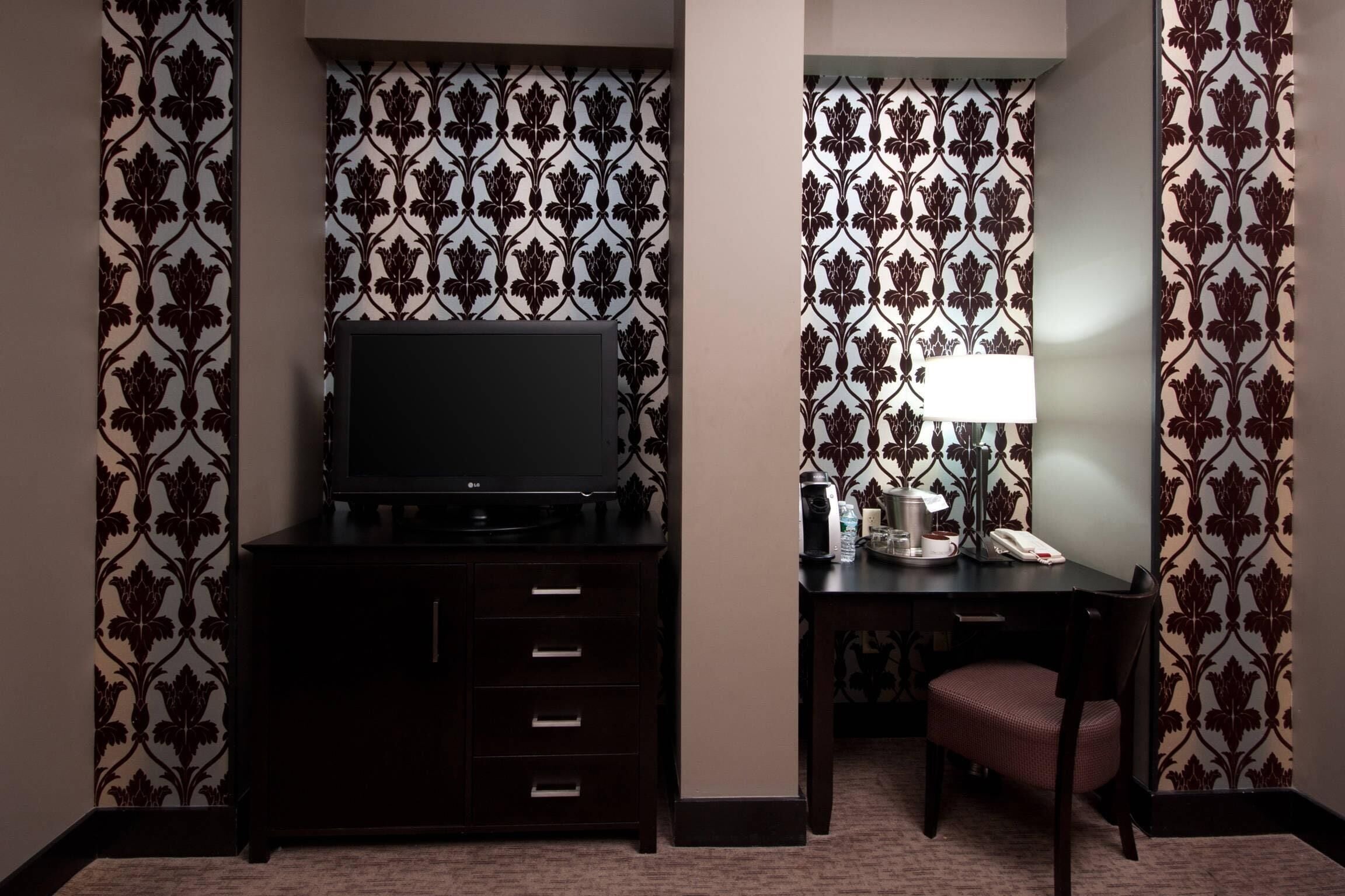 A hotel room with a TV on a dark wooden cabinet, a desk with a lamp, and a chair.