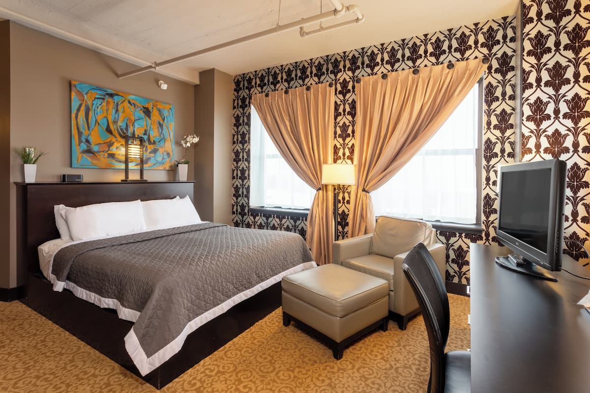 A hotel room with a bed, a desk with a television, a chair, a lamp, and a painting on the wall.