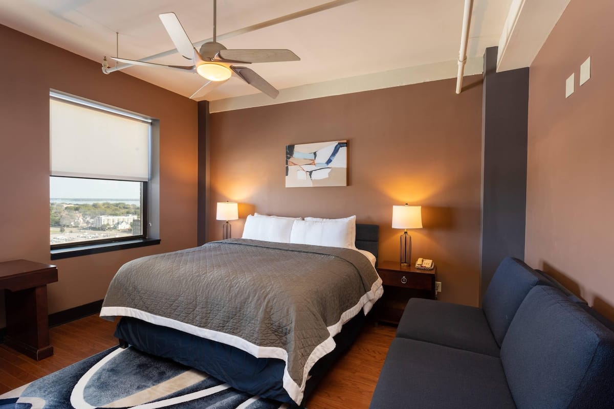 A hotel room with a bed, a ceiling fan, a nightstand with a lamp, a sofa, and a window with a view.