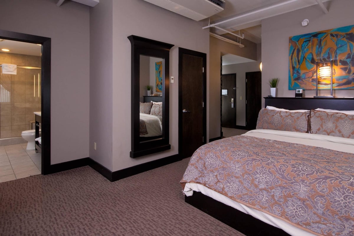 A hotel room with a large bed, a mirror, a painting on the wall, and a bathroom visible through an open door.