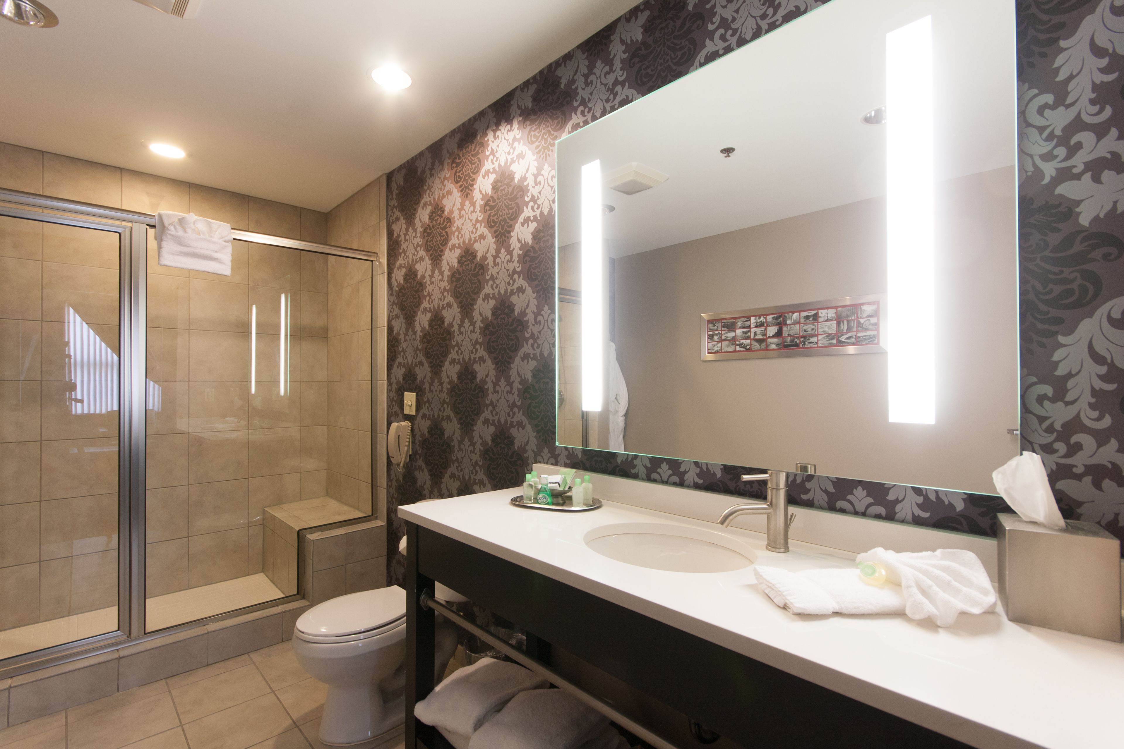 Un baño moderno con cabina de ducha de vidrio, inodoro blanco, espejo grande y tocador con lavamanos y toallero.