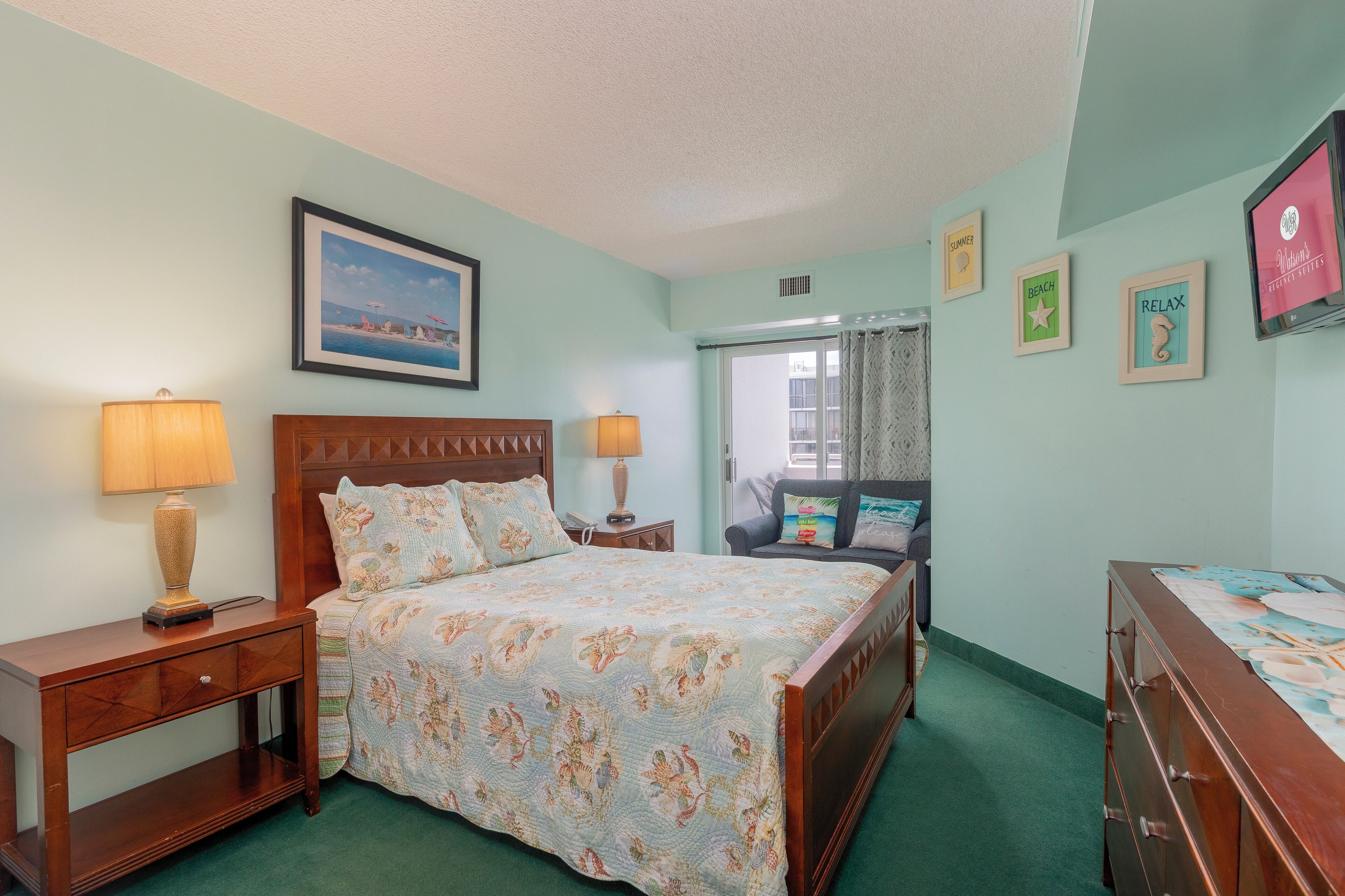 Une chambre à coucher comprenant un lit, deux tables de chevet avec des lampes, une commode, une fenêtre avec des rideaux et des éléments de décoration murale, notamment des tableaux encadrés et un téléviseur.