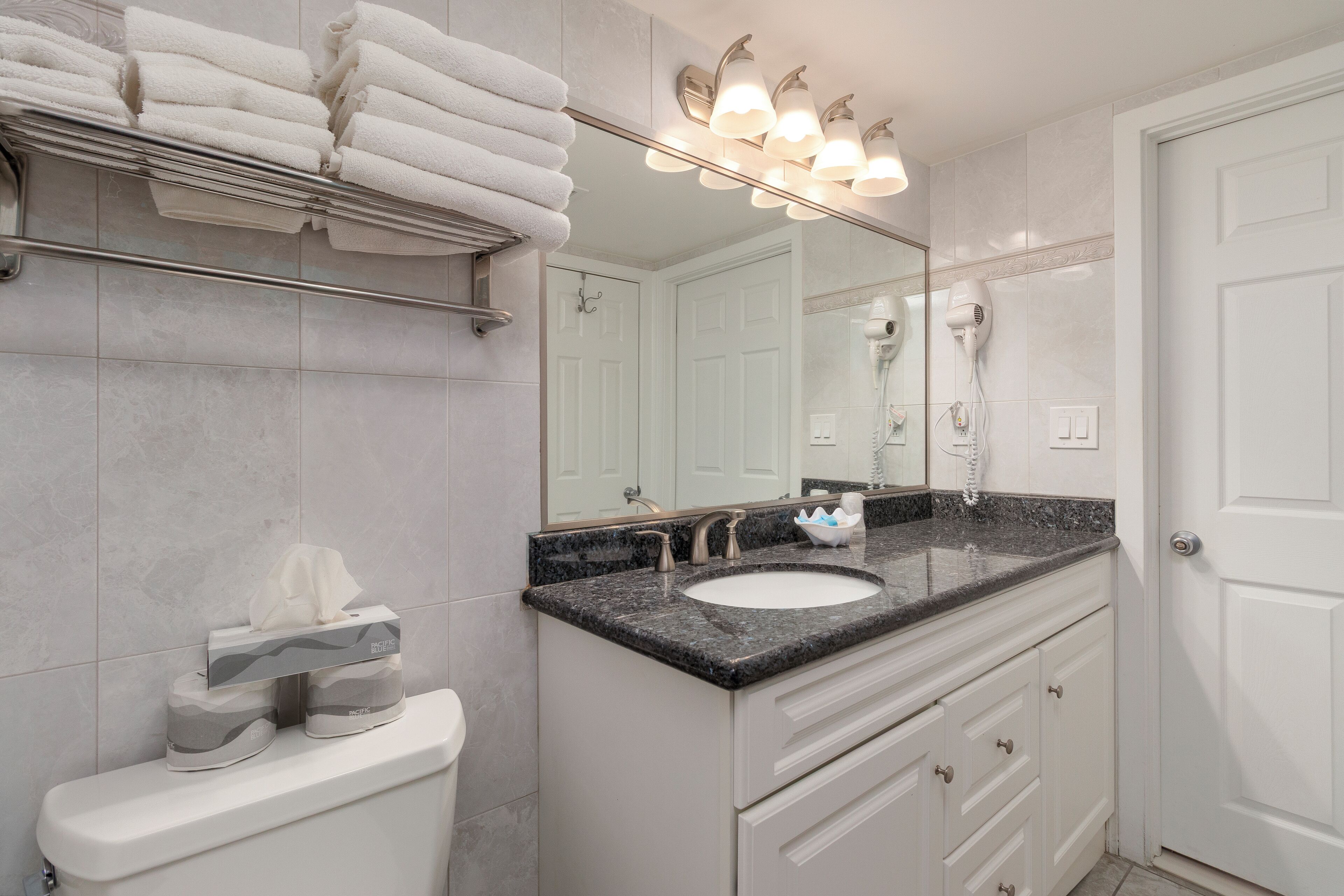 A bathroom with a white toilet, a sink with a granite countertop, a towel rack with folded towels, a mirror with lights, and a hairdryer on the wall.