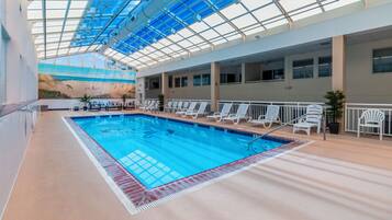 An indoor swimming pool with a glass ceiling, white lounge chairs, and a mural on the wall.