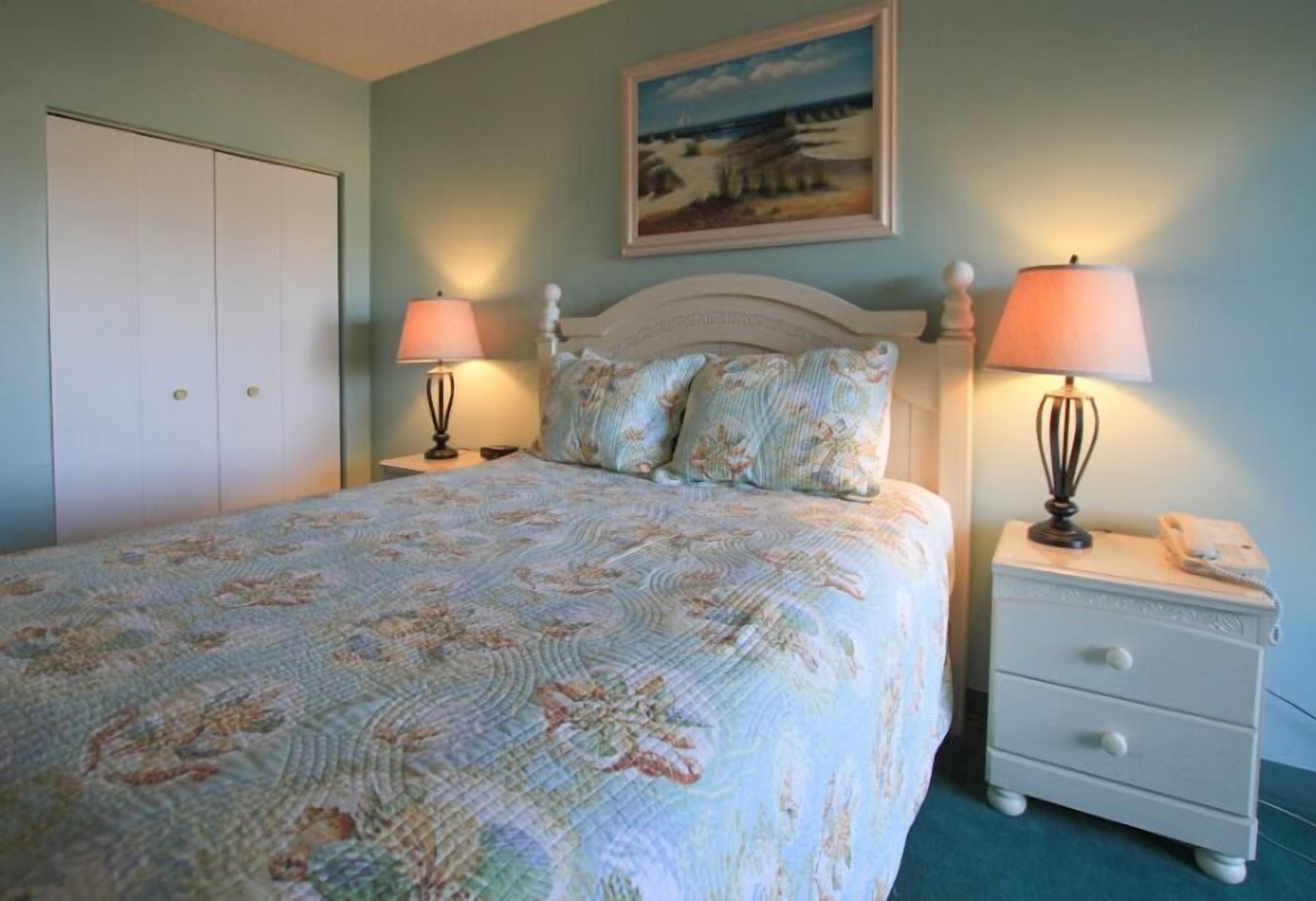 A bedroom with a bed, two bedside tables, a nightstand, a painting on the wall, and a door.