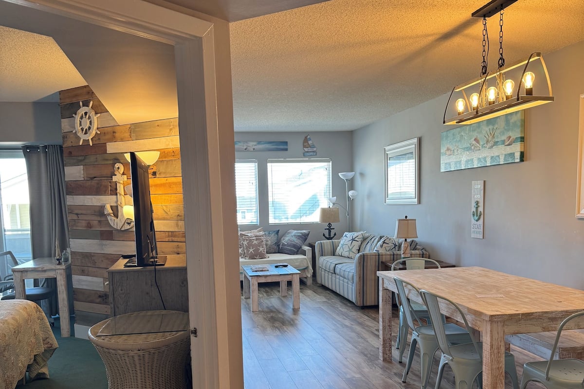 A spacious living area with a wooden floor, a dining table with chairs, a sofa, and a wicker chair.