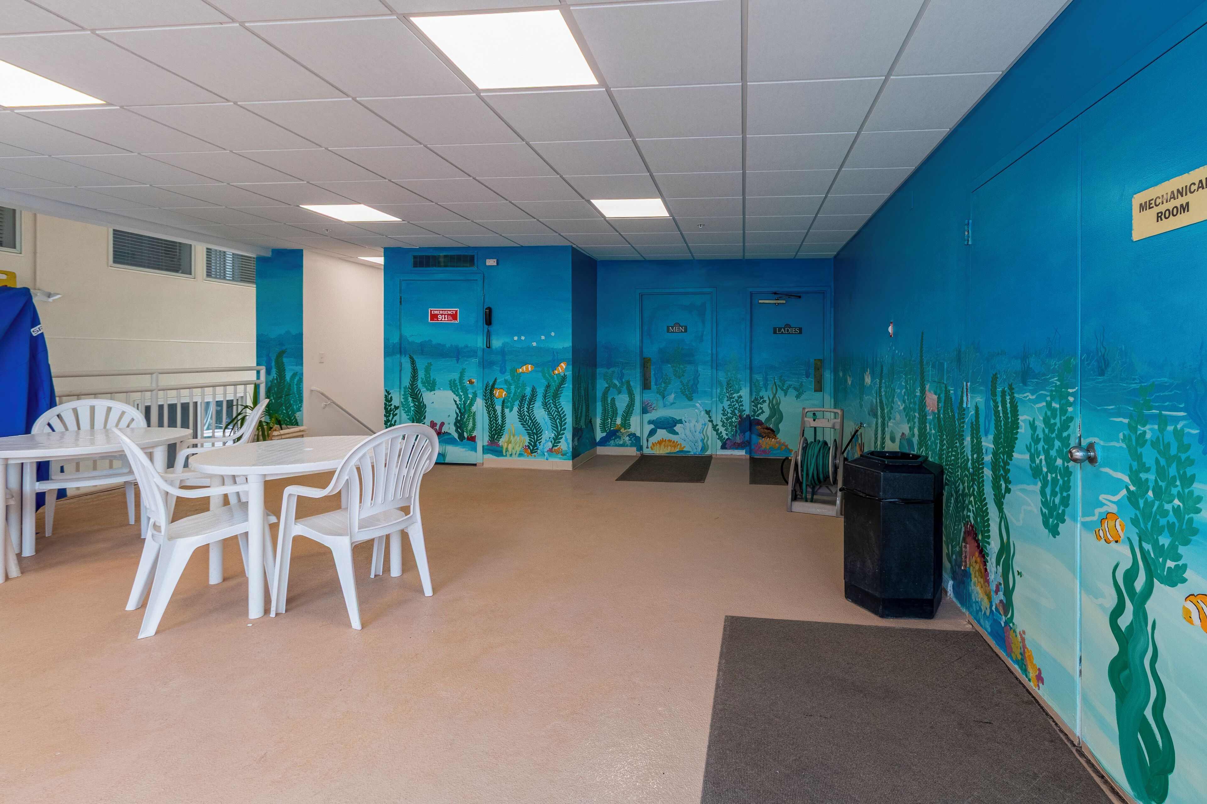 A room with a blue mural depicting an underwater scene, white plastic chairs, and tables. There are doors labeled "MEN" and "LADIES."