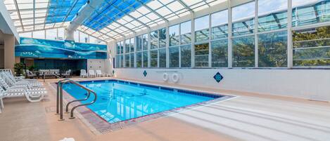 An indoor swimming pool with clear glass ceilings, lounge chairs, and a view of trees outside.