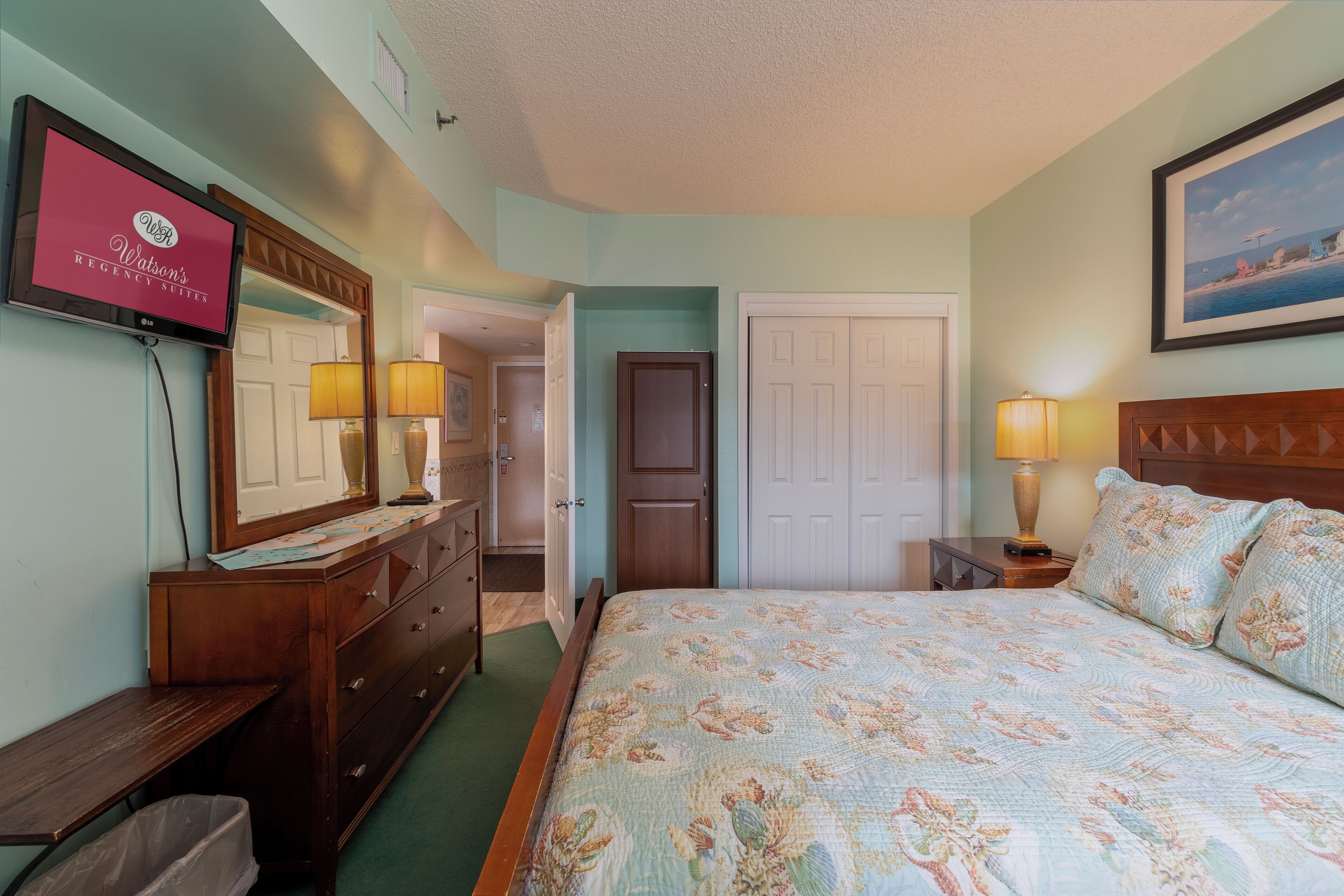 A bedroom with a bed, dresser, TV, and a door leading to another room.