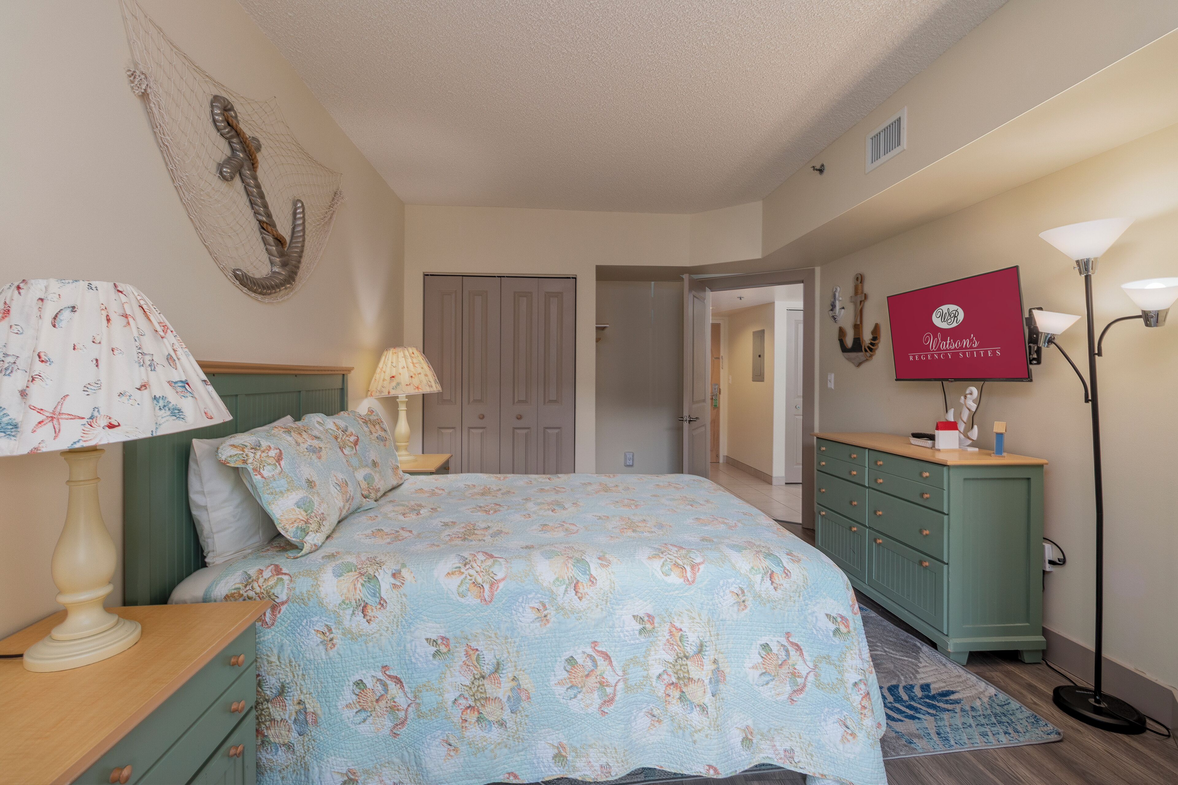 A bedroom with a bed, a nightstand, a lamp, a dresser, and a TV on the wall.