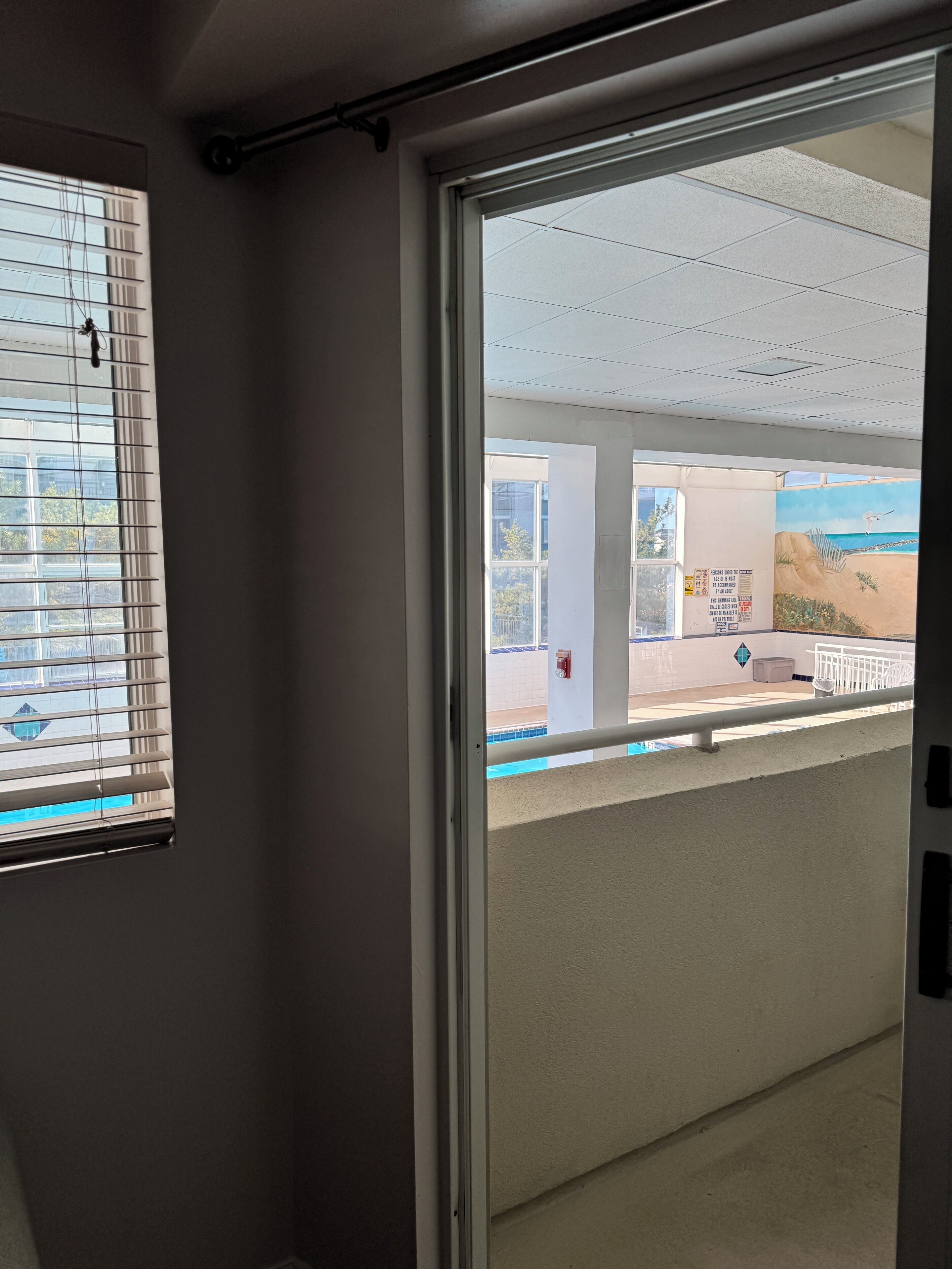 A room with a sliding glass door leading to a balcony with a view of a pool and beach.